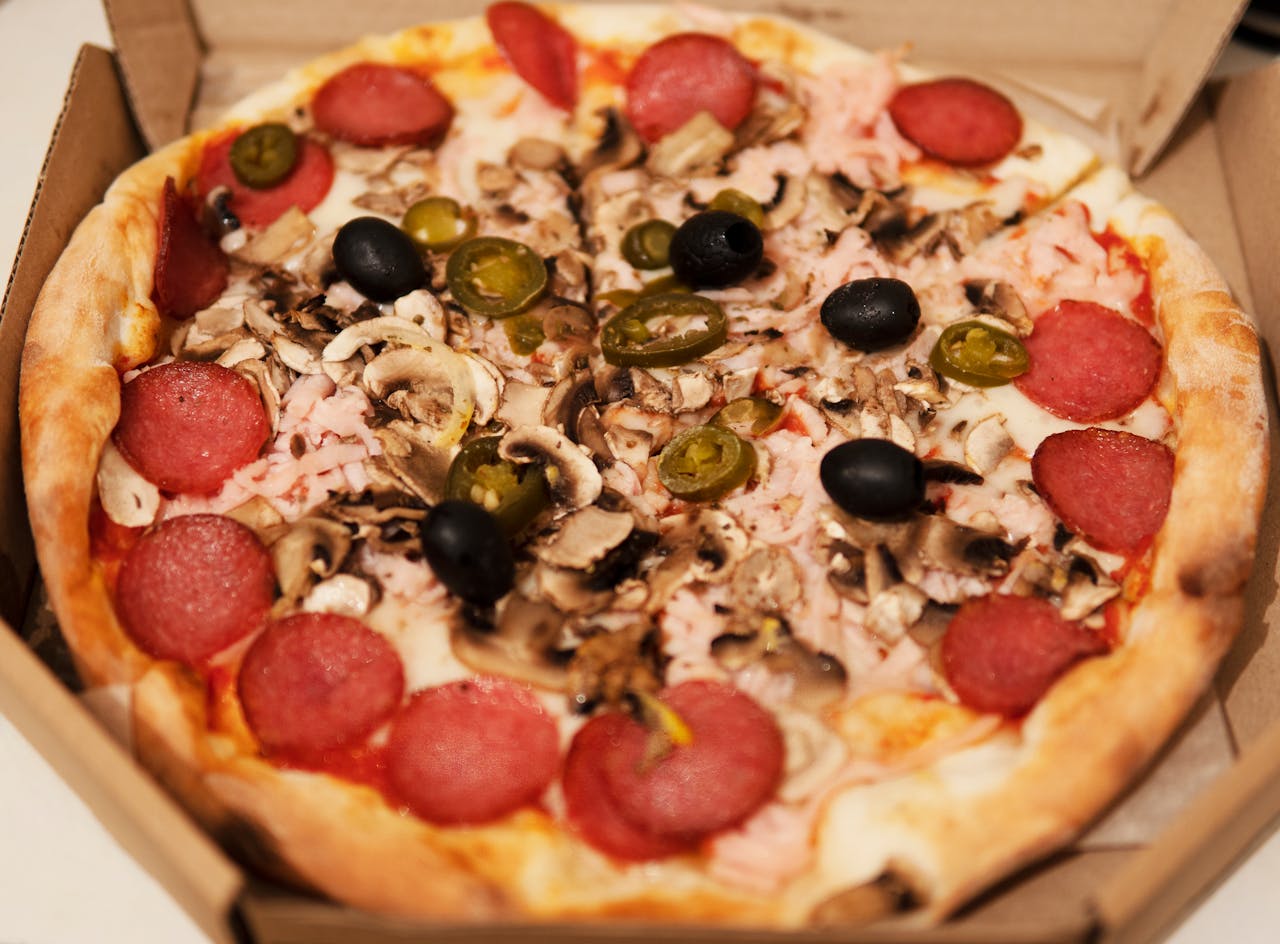 portfolio-01 Close-up of a freshly baked pizza topped with pepperoni, mushrooms, olives, and jalapenos in a cardboard box.